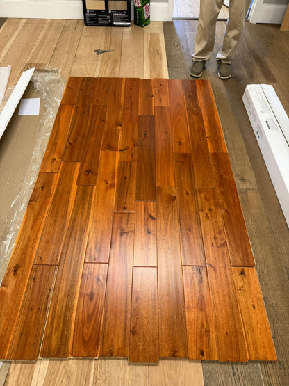 5" x 3/4" Solid Acacia Golden Hour Hardwood Flooring. Shown on a floor as a whole box. A person stands to the back right. 