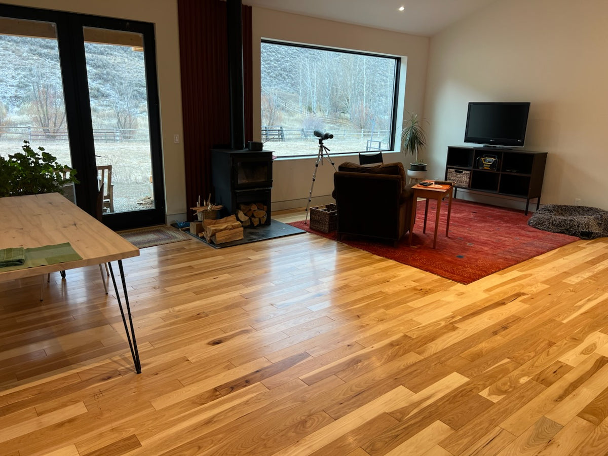 4 x 34 American Hickory Natural Hardwood Flooring. shown in a living room. There is a big window at center with a red rug in the middle of the room. Some furniture sits around the rug.