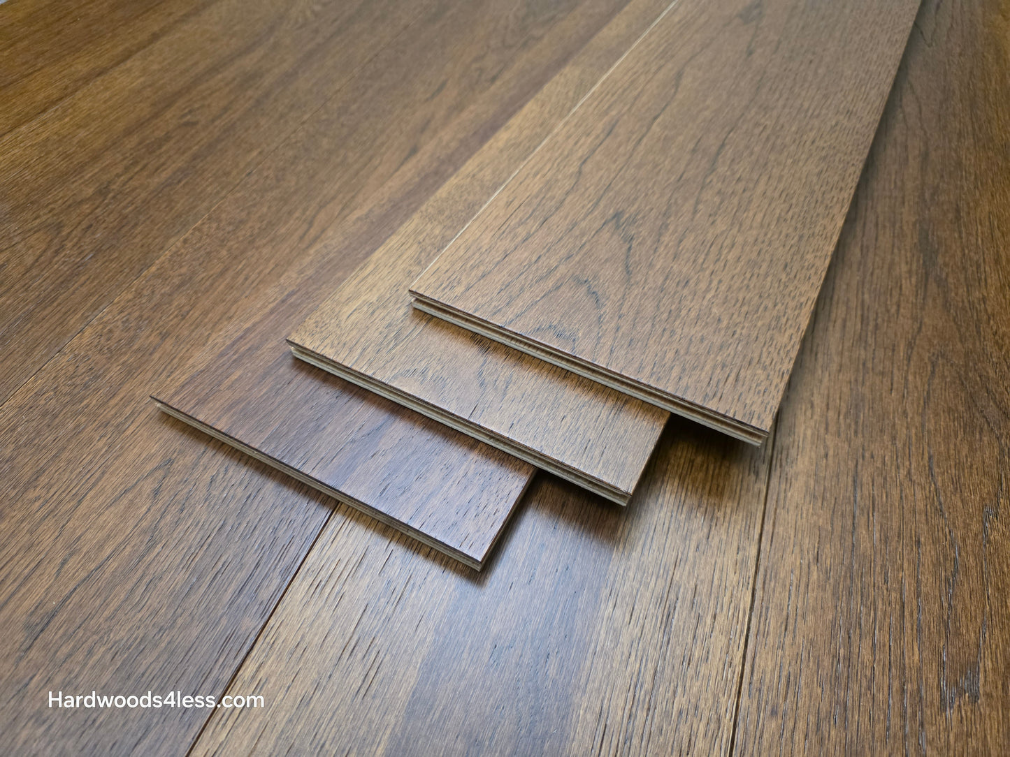 Close-up of 7 1/2" x 9/16" Engineered Hickory Alabay Hardwood Flooring stacked on top of the floor, with a visible brand name 'Hardwoods4less.com'.