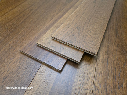 Close-up of 7 1/2" x 9/16" Engineered Hickory Alabay Hardwood Flooring stacked on top of the floor, with a visible brand name 'Hardwoods4less.com'.