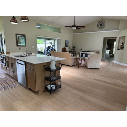 7 x 58 Engineered Red Oak Hudson Hardwood Flooring installed in a living room. There is a seating area, grey couch and seat in the center right. To the left side of the image there is a wooden island with a marble counter top and sink.
