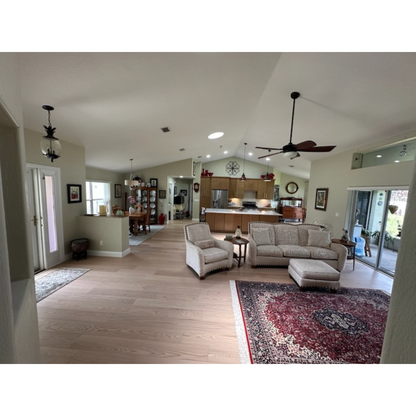 7 x 58 Engineered Red Oak Hudson Hardwood Flooring installed in a living room and kitchen space. There is a red and tan rug on the right side of the image. At center is a seating area, grey couch and seat. In the background is the kitchen featuring wooden cabinets. 