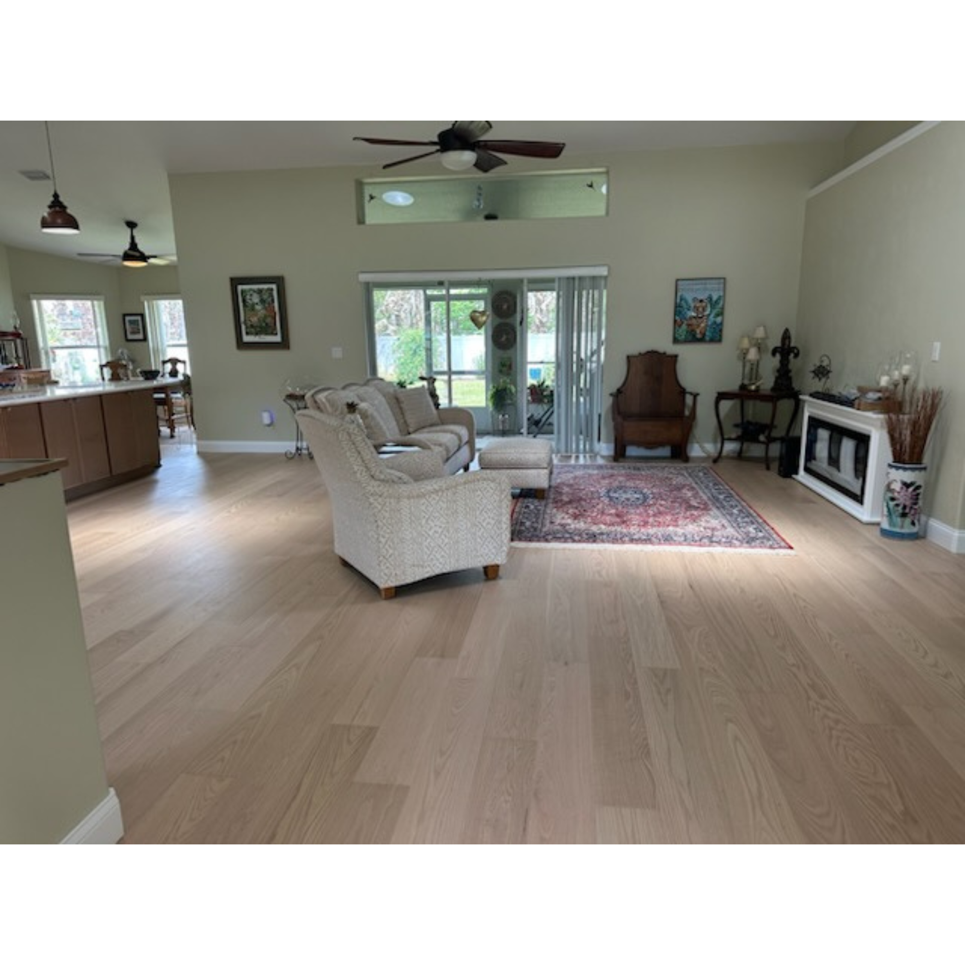 7 x 58 Engineered Red Oak Hudson Hardwood Flooring installed in a living room. There is a red and tan rug center right of the image. At center is a seating area, grey couch and seat. To the left side of the image you can see the start of the wooden cabinets.