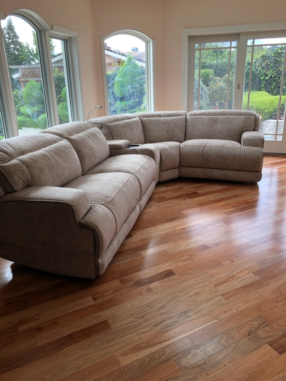 African Amendoim Hardwood Flooring. Installed in a living room with a tan couch in the center of the frame. Windows in the background show plants.